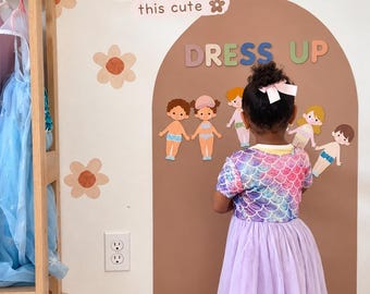 May include: A wall decal set featuring a brown arch, the text DRESS UP, and various paper doll figures. The wall also displays the text kind of chick to... make playtime this cute, surrounded by orange and brown flower decals on a white wall.