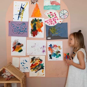 May include: A large, arched, peach-colored wall display board covered with various children's drawings, paintings, and photographs. The artwork includes colorful abstract paintings, a butterfly drawing, and a paper plate craft.