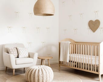 May include: A neutral-toned nursery with a wooden crib, armchair, and round rug. The room features a woven pendant light, a heart-shaped wall hanging, and a patterned wallpaper with bow designs. A knitted ottoman and small side table complete the space.