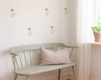 May include: A light green bench with a beige pillow sits in a corner with a white wall decorated with duck decals. A window with white curtains and a potted plant are visible in the background. The floor is made of wood.