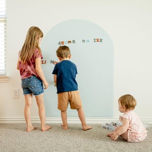 May include: A large, pale blue, arch-shaped wall decal applied to a white wall. The decal features a smooth surface where various colorful, small, magnetic-style alphabet letters are attached, including the characters A, B, C, M, R, N, I, X, and J.