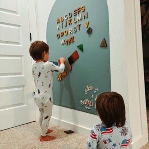 May include: A large, arched, sage green magnetic wall board featuring colorful wooden alphabet letters from A to Z arranged at the top, along with various geometric shapes and small decorative magnets scattered across the surface.