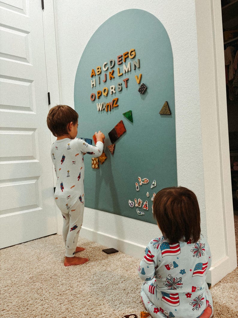 May include: A large, arched, sage green magnetic wall board featuring colorful wooden alphabet letters from A to Z arranged at the top, along with various geometric shapes and small decorative magnets scattered across the surface.