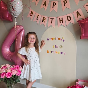 May include: A large, arched, beige wooden backdrop board featuring colorful letter stickers that read "Angelina's 4th Birthday" with a smiley face icon. The board is decorated with pink "Happy Birthday" bunting, a Minnie Mouse balloon, and a large number 4 balloon.