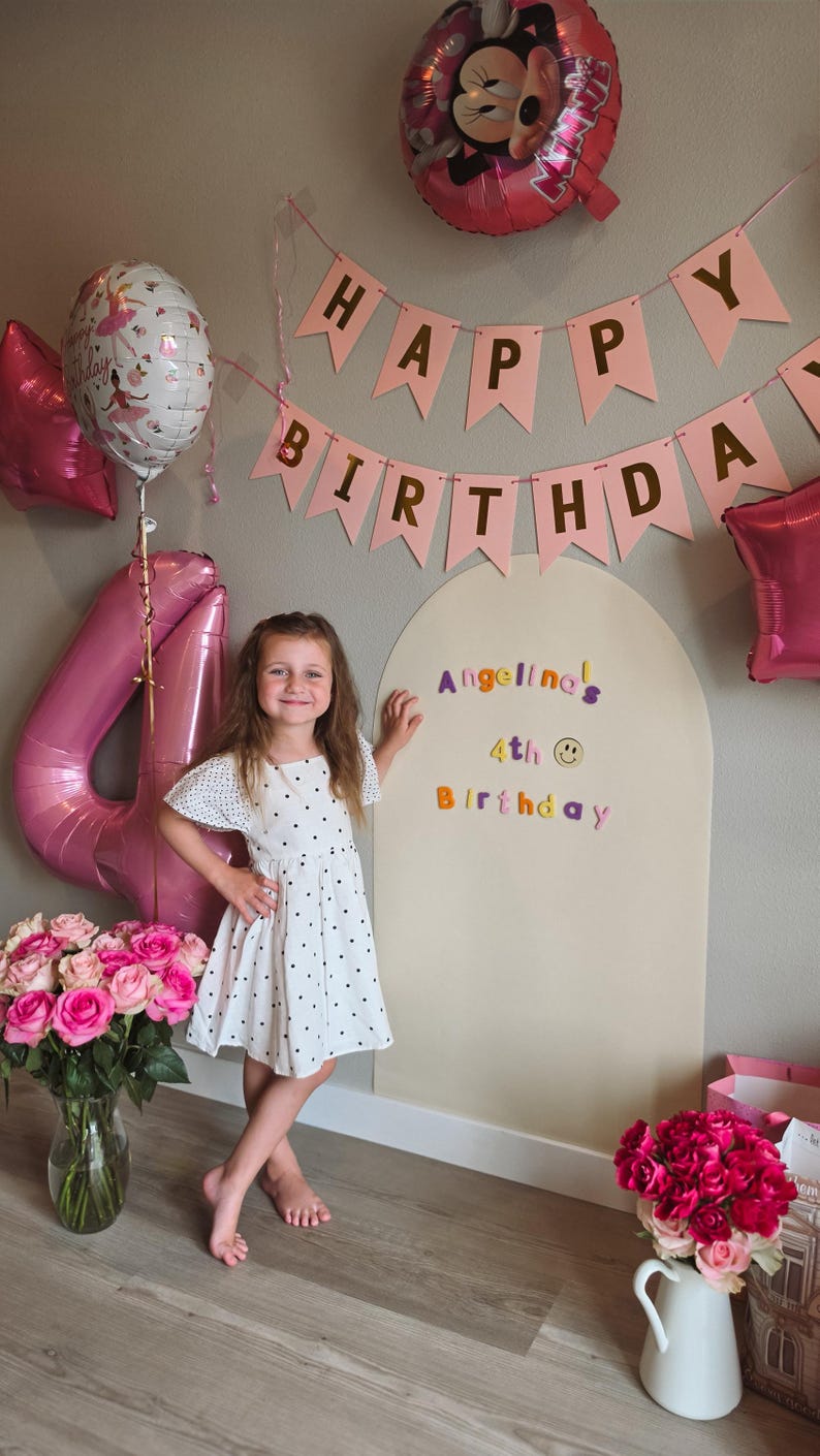May include: A large, arched, beige wooden backdrop board featuring colorful letter stickers that read "Angelina's 4th Birthday" with a smiley face icon. The board is decorated with pink "Happy Birthday" bunting, a Minnie Mouse balloon, and a large number 4 balloon.