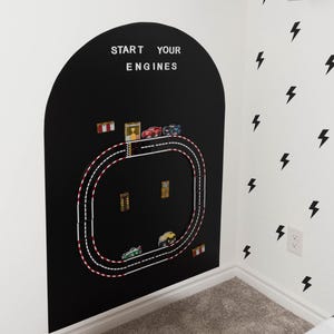 May include: A black arched wall decal featuring a white race track graphic with the text START YOUR ENGINES at the top. The track includes small toy cars placed along the path, with red and white checkered accents and various small racing icons.