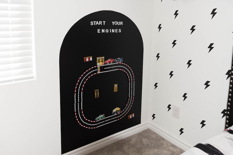 May include: A black arched wall decal featuring a white race track graphic with the text START YOUR ENGINES at the top. The track includes small toy cars placed along the path, with red and white checkered accents and various small racing icons.