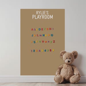 May include: A tan wall decal featuring the white text RYLIE'S PLAYROOM above a colorful alphabet and the numbers 1 through 9. A light brown plush teddy bear sits on the floor in front of the decal, which is applied to a plain wall.