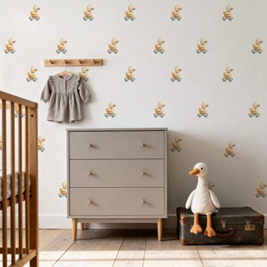 May include: A nursery scene with a light gray dresser, a crib, and a stuffed goose. The wall is decorated with yellow duck decals on wheels. A gray dress hangs on a wooden rack. The room has a warm, inviting atmosphere.