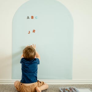 May include: A large, light blue, arch-shaped wall decal applied to a wall. The decal features magnetic properties, with several small, colorful, individual letter magnets attached to its surface, including the letters A, B, C, J, and R.