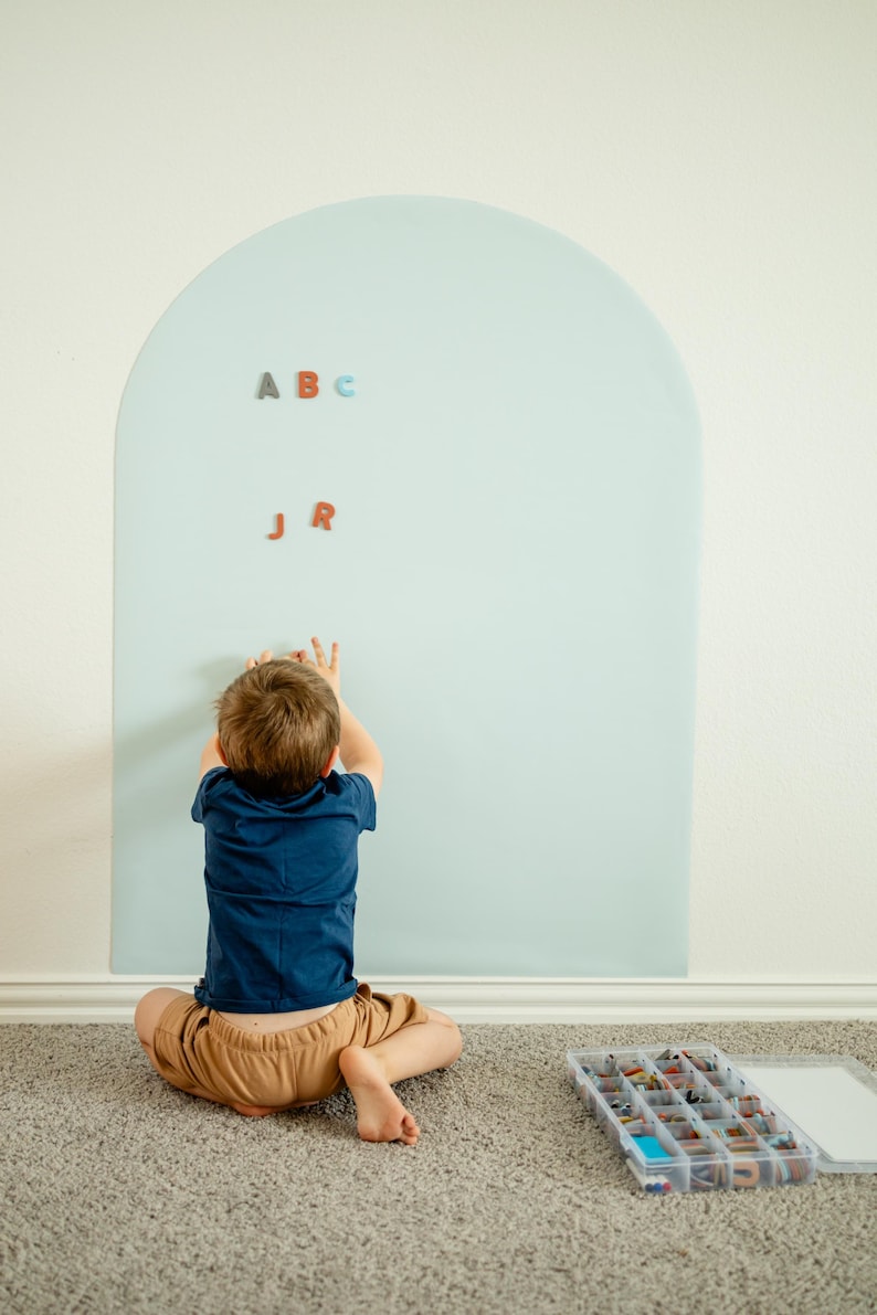 May include: A large, light blue, arch-shaped wall decal applied to a wall. The decal features magnetic properties, with several small, colorful, individual letter magnets attached to its surface, including the letters A, B, C, J, and R.