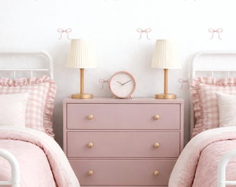 May include: A bedroom scene featuring two white metal bed frames with pink gingham ruffled bedding and matching quilts. Between the beds sits a dusty pink three-drawer dresser with gold knobs, two cream lamps, and a round pink analog clock.