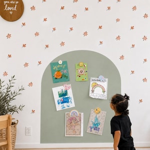May include: A sage green arch painted on a white wall decorated with small orange flower decals. The arch serves as a display area for five rectangular papers held by various colorful magnets, including a donut, sun, and rainbow shape.