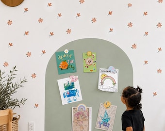 May include: A sage green arch painted on a white wall decorated with small orange flower decals. The arch serves as a display area for five rectangular papers held by various colorful magnets, including a donut, sun, and rainbow shape.