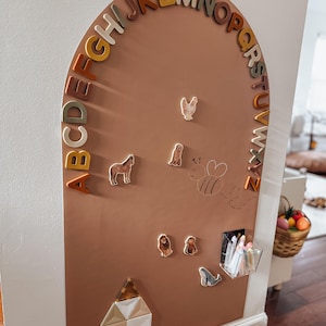 May include: An arched, terracotta-colored wall board featuring the alphabet in raised, multi-colored letters along the curve. It includes magnetic animal figures, a line-drawn bee, a clear acrylic storage bin with markers, and geometric shapes.