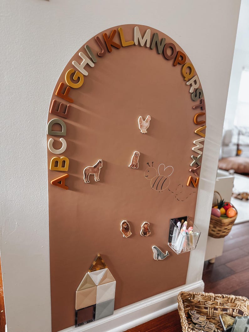 May include: An arched, terracotta-colored wall board featuring the alphabet in raised, multi-colored letters along the curve. It includes magnetic animal figures, a line-drawn bee, a clear acrylic storage bin with markers, and geometric shapes.