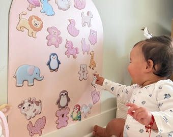 May include: An arched, light pink wall display board featuring various colorful animal stickers, including a flamingo, lion, elephant, penguin, sloth, and koala, arranged across the surface for interactive play and visual engagement.