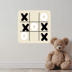 May include: A beige wall decal featuring a tic-tac-toe game with black X's and white O's. A brown teddy bear sits to the right of the decal. The game board is a square with rounded corners.