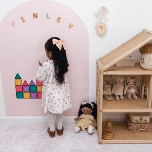May include: A large pink arch wall decal with the name FINLEY in beige letters, featuring colorful magnetic building tiles attached to the surface. A rattan dollhouse with three shelves holds plush toys, and a fabric bunting hangs above.