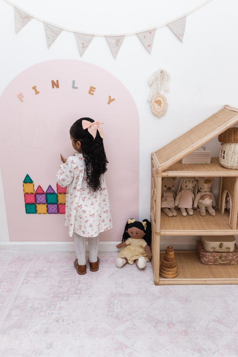 May include: A large pink arch wall decal with the name FINLEY in beige letters, featuring colorful magnetic building tiles attached to the surface. A rattan dollhouse with three shelves holds plush toys, and a fabric bunting hangs above.