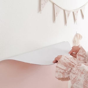 May include: A large, smooth, pale pink adhesive wall decal being applied to a white wall. Above the decal, a string of small, triangular, off-white fabric pennant flags with delicate floral embroidery hangs horizontally against the wall surface.