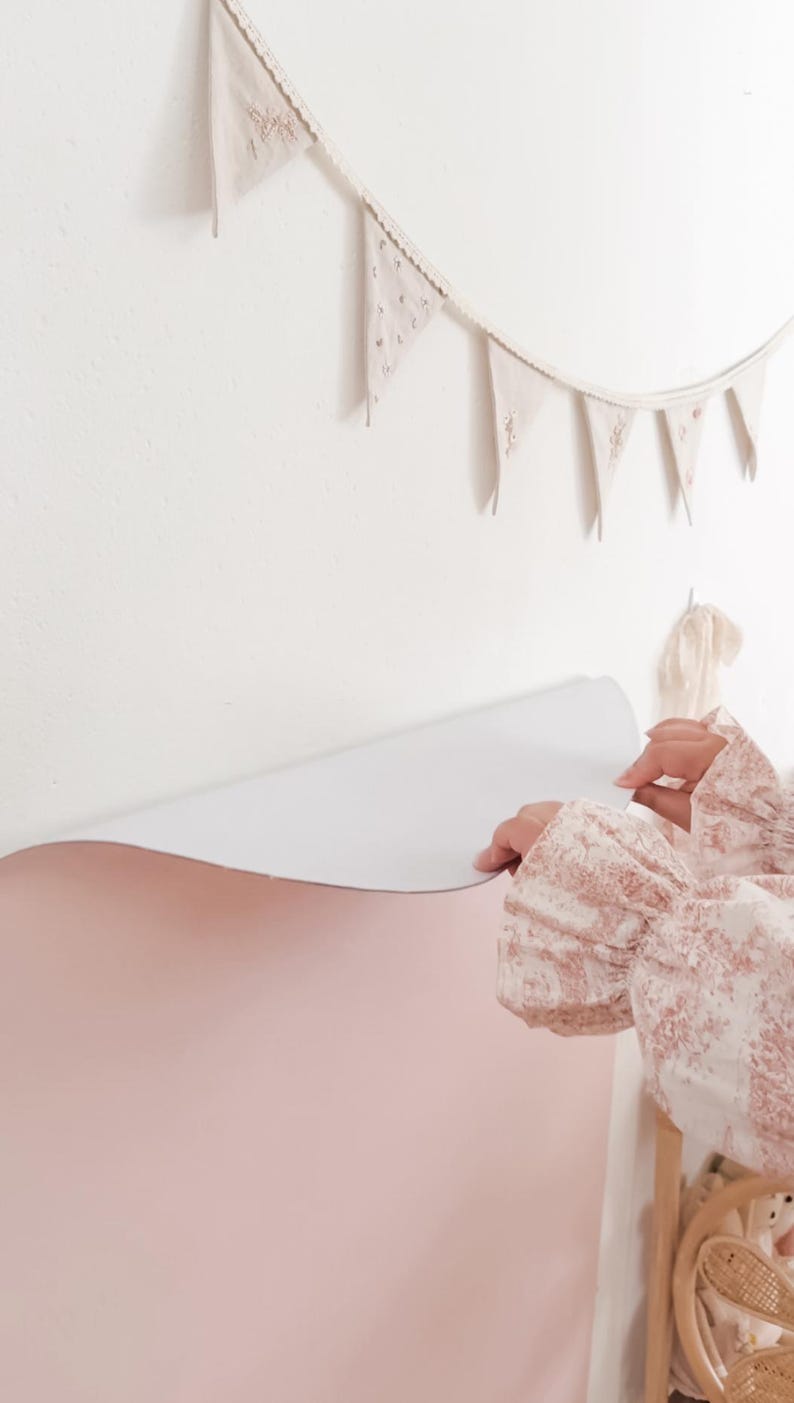 May include: A large, smooth, pale pink adhesive wall decal being applied to a white wall. Above the decal, a string of small, triangular, off-white fabric pennant flags with delicate floral embroidery hangs horizontally against the wall surface.
