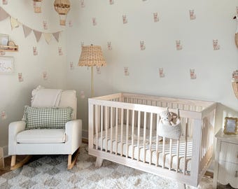 May include: A nursery with a light color palette. The room features a crib, a rocking chair, and a fluffy rug. The walls are decorated with cat decals and wicker hot air balloons. A woven lamp and a small nightstand complete the room.