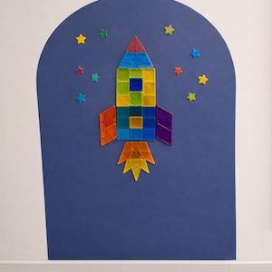 May include: A rocket ship constructed from colorful translucent magnetic tiles, featuring a red nose cone, a blue, green, and yellow body, orange and purple fins, and a yellow and orange flame, surrounded by small star-shaped magnetic tiles.