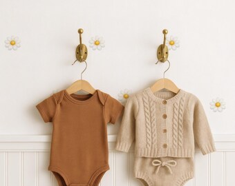 May include: Two baby outfits hanging on brass hooks against a white wall with daisy decals. One is a brown short-sleeved bodysuit. The other is a beige cable-knit cardigan and matching shorts set.
