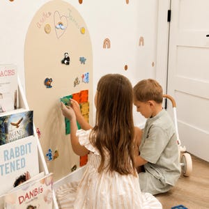 May include: A large, beige, arch-shaped magnetic wall board featuring a hand-drawn heart with the names "a selle" and "James" written inside. The board is decorated with various small magnetic figures, including a panda and a squirrel.
