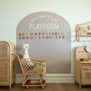 May include: A wall decal featuring a mauve arch shape with the white text CORA, EMMA & LILA'S PLAYROOM and a full alphabet in multi-colored letters. The decal is displayed on a wall behind a rattan toy shopping cart containing a white plush llama.