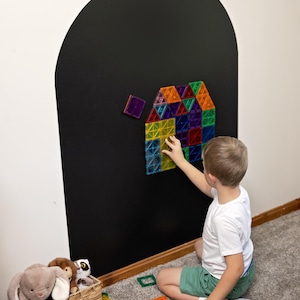 May include: A set of colorful, translucent magnetic building tiles arranged into a house shape on a large, black, arch-shaped wall decal. The tiles are square and triangular in shades of red, blue, green, yellow, orange, and purple.
