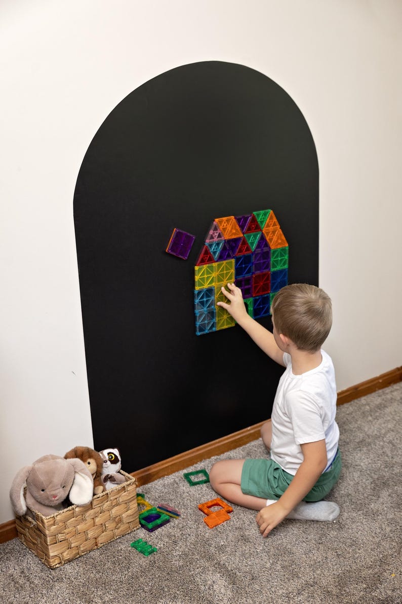 May include: A set of colorful, translucent magnetic building tiles arranged into a house shape on a large, black, arch-shaped wall decal. The tiles are square and triangular in shades of red, blue, green, yellow, orange, and purple.