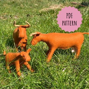 May include: A PDF pattern for sewing three felt cows. The cows are orange with white markings and are standing in a grassy field. The pattern is available for download.