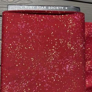 Op de afbeelding: Een gevouwen stuk dieprode stof met een gespikkeld patroon van kleine gele en roze stippen. De bovenrand van de stof is vastgezet met een zwart label met de witte tekst RUBY STAR SOCIETY gedrukt tussen twee witte sterren.