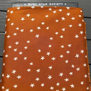 May include: A folded piece of fabric featuring a burnt orange background with a repeating pattern of small white stars. The top edge of the fabric shows a black label with the white text RUBY STAR SOCIETY printed between two white stars.