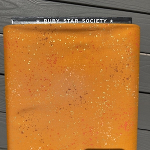 Peut inclure: Un morceau de tissu plié d'une teinte jaune moutarde vibrante, présentant un motif tacheté de petits points orange et blancs sur la surface. Le bord supérieur affiche une étiquette noire avec le texte blanc Ruby Star Society imprimé dessus.