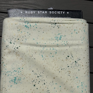 May include: A folded piece of off-white fabric featuring a speckled pattern of teal and metallic gold dots. The fabric is topped with a black label that reads RUBY STAR SOCIETY in white capital letters with a star on each side of the text.