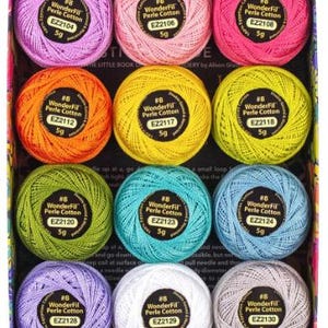 Op de afbeelding: Een doos met kleurrijke WonderFil Perle Cotton garenbollen. De collectie bevat tinten roze, oranje, geel, groen, blauw, paars, wit en grijs. Elke bol is gemarkeerd met het garentype en gewicht, 5g.