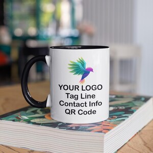 May include: White ceramic mug with a black rim and handle. The mug features a colorful bird logo, the text "YOUR LOGO", "Tag Line", "Contact Info", and "QR Code". The mug is sitting on a stack of books.