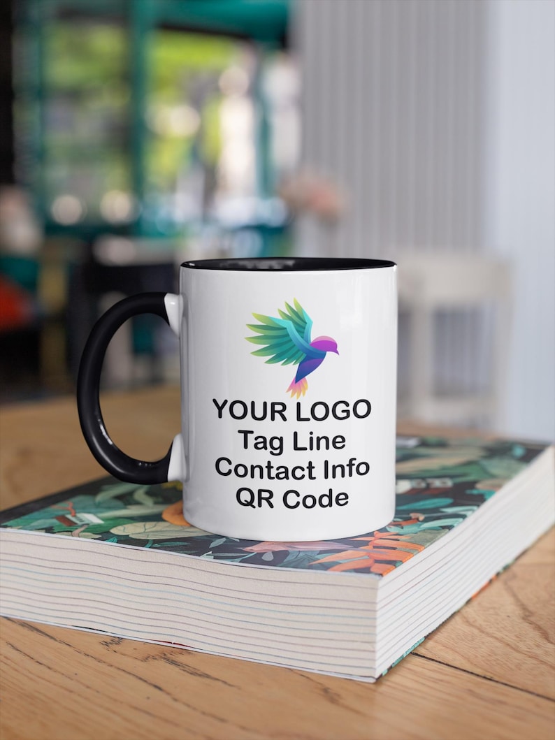 May include: White ceramic mug with a black rim and handle. The mug features a colorful bird logo, the text "YOUR LOGO", "Tag Line", "Contact Info", and "QR Code". The mug is sitting on a stack of books.
