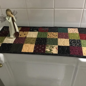 May include: A rectangular patchwork quilt runner featuring a variety of fabric squares in shades of green, burgundy, black, and cream with floral and star patterns, finished with a black border, and a small angel figurine with wire wings.