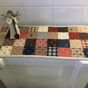 May include: A patchwork toilet tank cover with a variety of patterned squares in shades of red, blue, brown, and cream. A small angel figurine with a shovel stands on the cover. The cover is rectangular and edged with a cream-colored border.