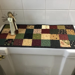 May include: A decorative quilted toilet tank topper featuring a patchwork design in various colors like burgundy, green, and cream. The topper has a black border and is adorned with a small angel figurine. The overall theme is country or farmhouse style.