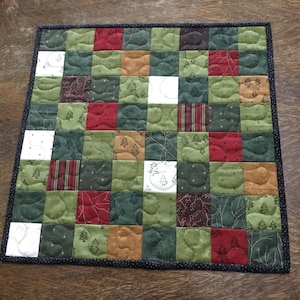 May include: A square patchwork quilt featuring a grid of fabric squares in shades of forest green, deep red, brown, and white. The fabric squares display various patterns including small pine trees, thin lines, and berries, finished with a dark border.