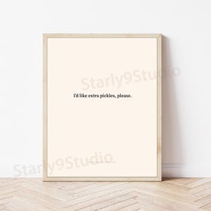 May include: A rectangular wall art print in a light wood frame featuring a cream-colored background with the black text "I'd like extra pickles, please." centered in the middle of the print. The design is simple and clean with a light wood border.