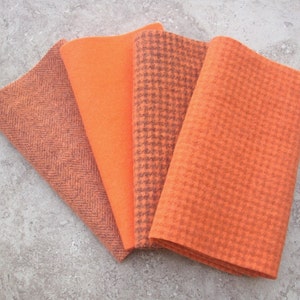 May include: Four orange fabric swatches in varying textures. The fabrics include a herringbone weave, a solid color, a houndstooth pattern, and a textured weave. The swatches are arranged in a fan shape, showcasing the different textures and shades of orange.