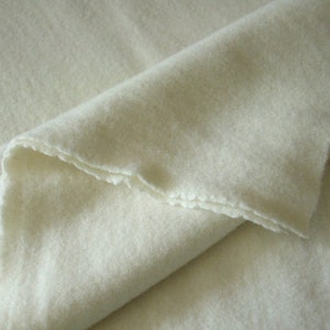 May include: A folded piece of off-white fabric with a soft, felted texture. The material features a dense, matte surface with visible fibers and raw, slightly uneven edges along the fold, showcasing a consistent light cream color throughout.