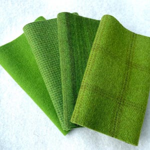 May include: Four folded fabric swatches in shades of green, arranged in a fan shape. The fabrics display varying textures, including a solid, a small houndstooth pattern, and a plaid design. The swatches are displayed on a white surface.