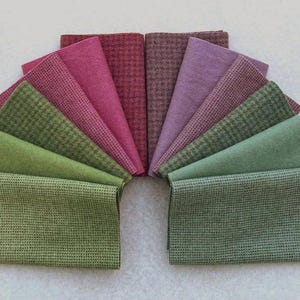 May include: A collection of fabric swatches arranged in a semi-circle. The swatches display a variety of colors, including shades of green, pink, and purple. Some swatches have a textured pattern, while others are solid. The fabrics are likely for sewing or crafting.